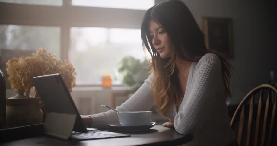Woman scrolls on a touchscreen of a laptop as she sits at a table with a bowl of food in front of her