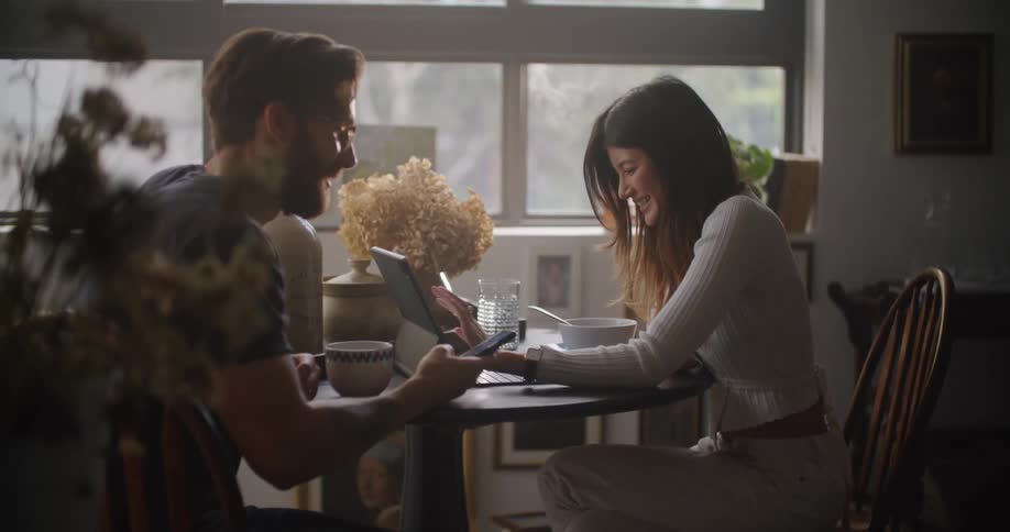 Man and woman sit at a table as the woman works on a laptop and the man shows her something on his cellphone
