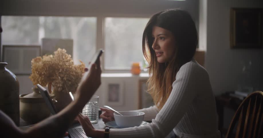 Man sitting at the table showing something on his smartphone to a woman that is using her laptop next to him