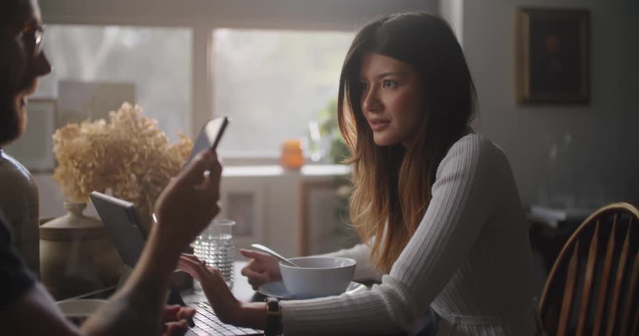 A man shows the screen of his smartphone to a woman sitting across from him at a dining table in a home kitchen