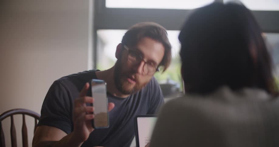 Man sitting at the table inside the house showing something on his smartphone to the woman sitting in front of him