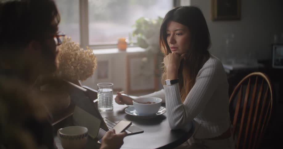A man uses his cell phone as he sits at a table and talks with a woman that is looking at a laptop