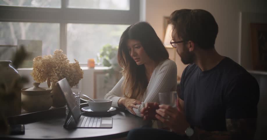 Man with glasses beard and black shirt holds glass of water sits next to woman with brown hair and white shirt using tablet at home