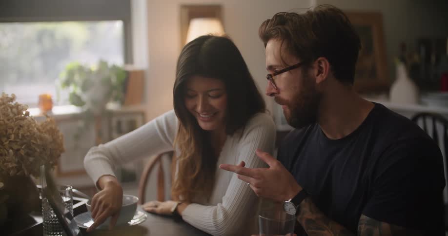 A woman points at objects on the screen of laptop while speaking to a man sitting next to her at a table in a house during the day