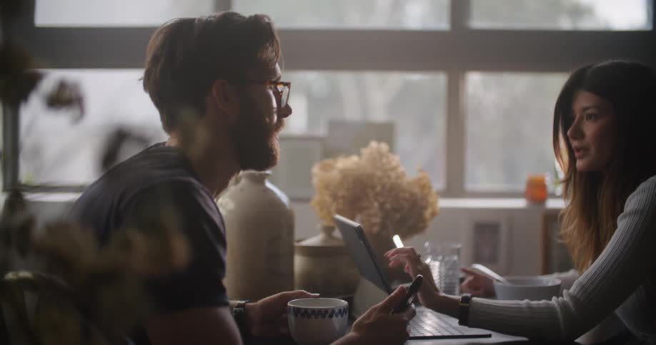 Man and woman sitting at the table using their smartphone and laptop while talking to each other