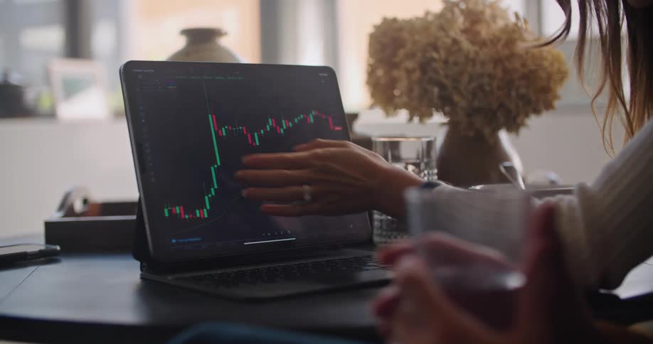 Woman sitting at the table inside the house analyzing and navigating through a candle chart on her laptop screen