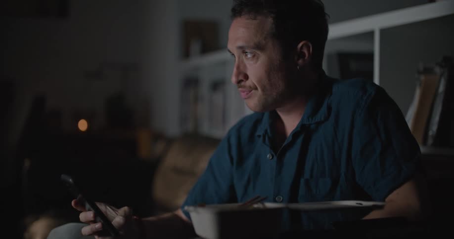 A man sits on a couch in front of his television with food while looking at his phone