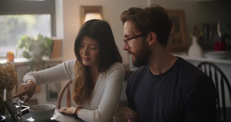 Woman sits next to a man at the table and explains something to him while pointing at the laptop screen