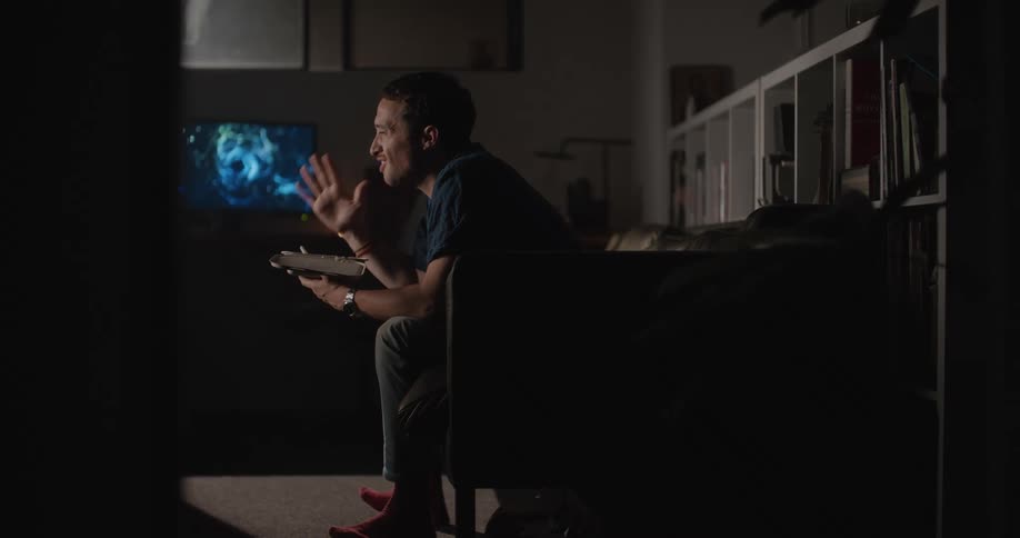 A man sits on a couch and excitedly holds up his hand while watching his television