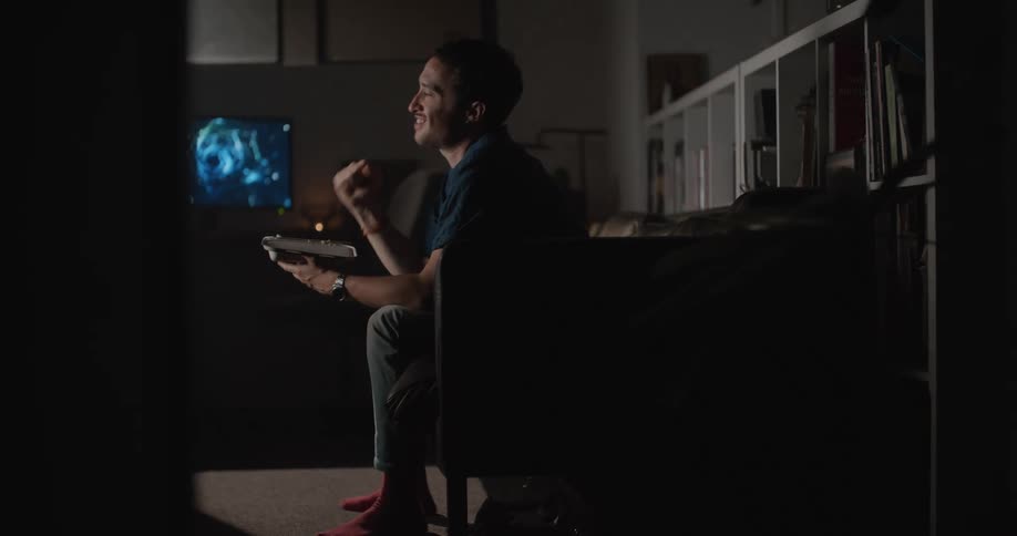An adult male sits on a couch and pumps his fist excitedly as he watches television