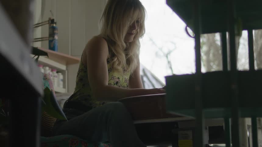 A blonde woman shaping clay while sitting at a pottery wheel