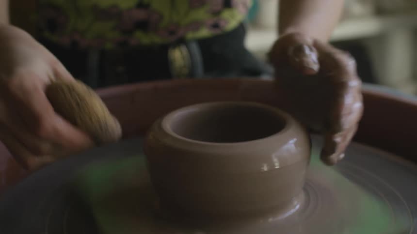 A woman uses a pottery wheel to sculpt clay indoors during the day