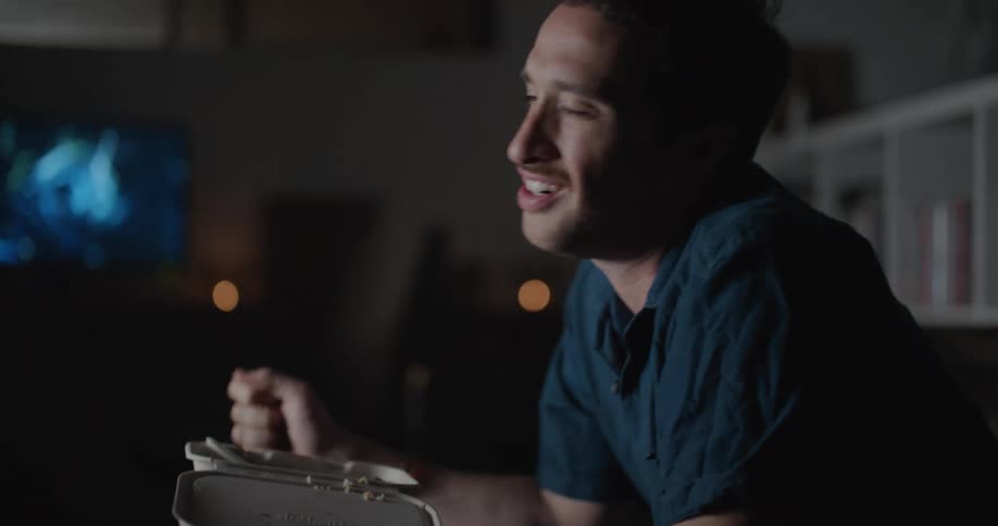 A man shaking his head in disappointment while eating takeout and watching television