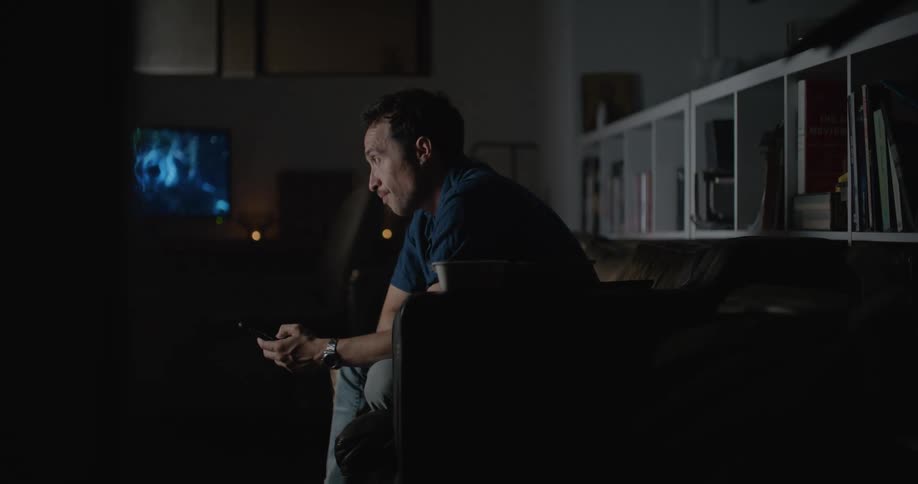 Man sitting on the couch at the living room scrolling through his smartphone while watching tv