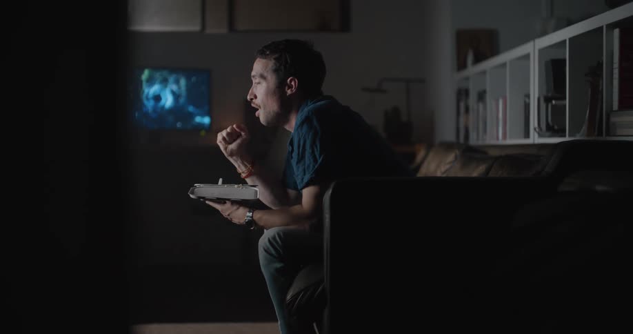 A man sitting on his couch at home holding a dinner container leans forward watching intently then pumps his fist in excitement