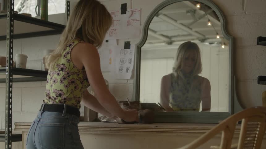 A woman kneads clay in front of a mirror in a workshop during the day