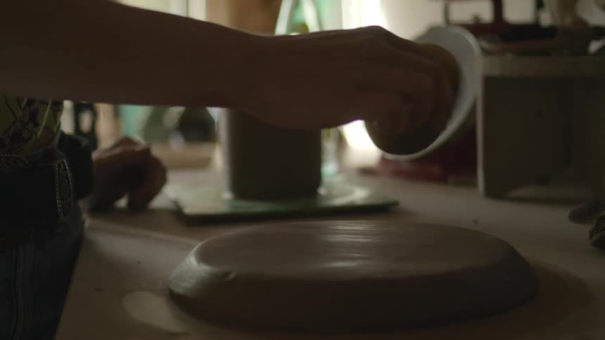 A woman keeps a flattened piece of clay moist by rubbing it with a wet sponge