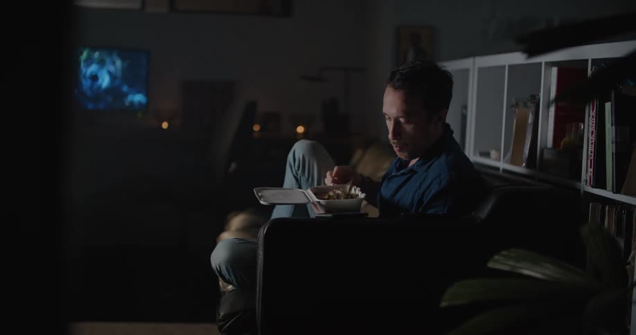 A man using his phone while eating takeout on a couch and watching a movie