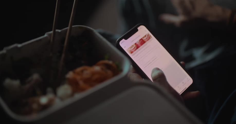 A person scrolling on the phone with some food on a plastic container next to it