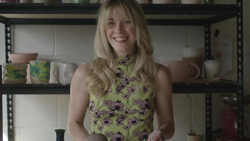 A woman standing in front of a shelf full of pottery smiles and tosses a ball of clay into the air