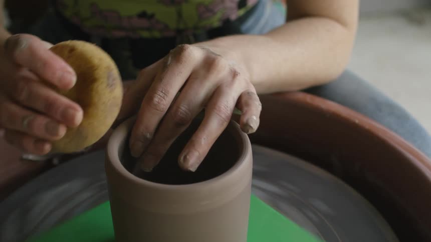 A woman carefully shapes a piece of clay that is spinning on a pottery wheel while keeping it wet with a sponge