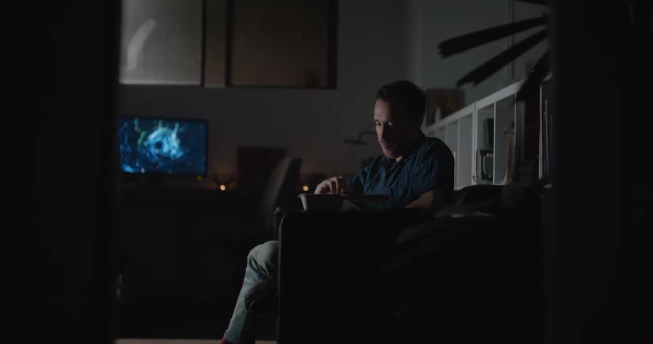 A man sits on a couch in front of his television while eating and looking at his phone