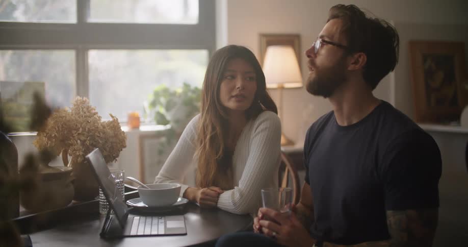 A man and woman gesture at the screen of a laptop while talking at a table