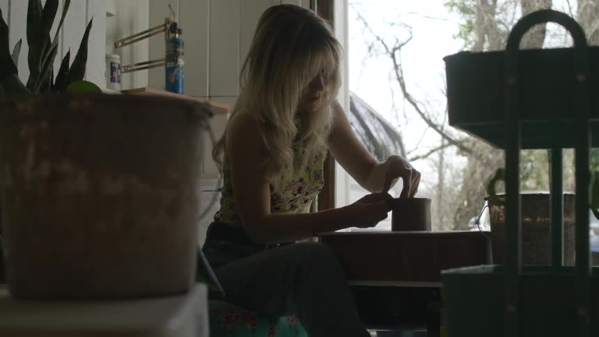 A woman with long blonde hair concentrates on shaping a piece of clay on her pottery wheel