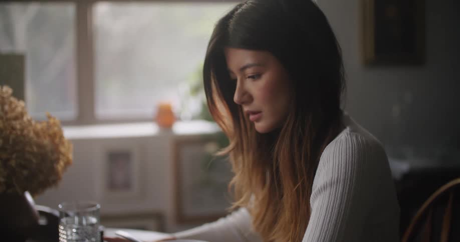Man uses smartphone next woman with brown straight hair and gray shirt working on tablet at table in the kitchen at home