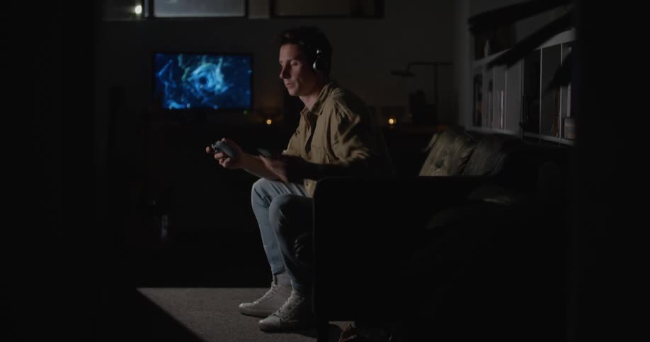 A man checks his smartphone before sitting it down and returning to playing a video game in the dark living room of a house at night