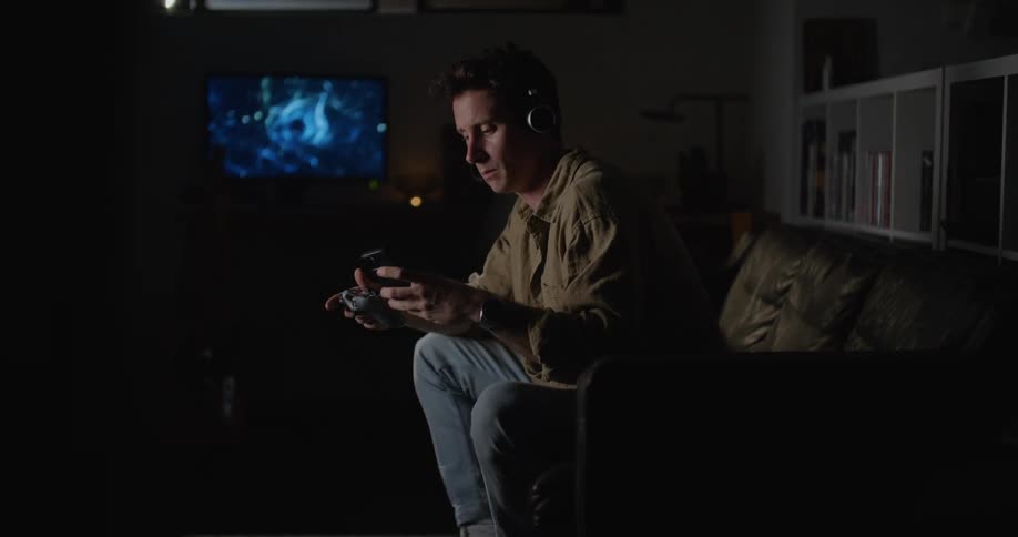 An adult male wearing head phones checks his cell phone while playing video games