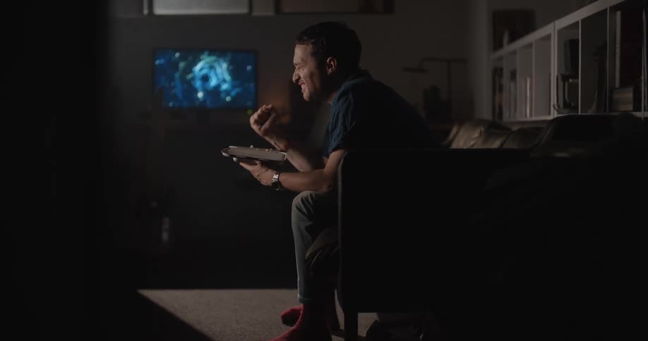 Man sitting on a sofa holding a container of food clenches his fist and looks upset as a television sits to his right