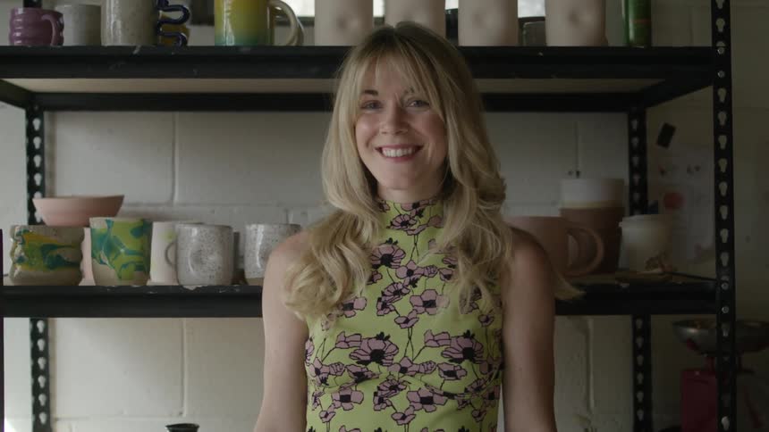 A woman standing in front of a shelves holding pottery smiles