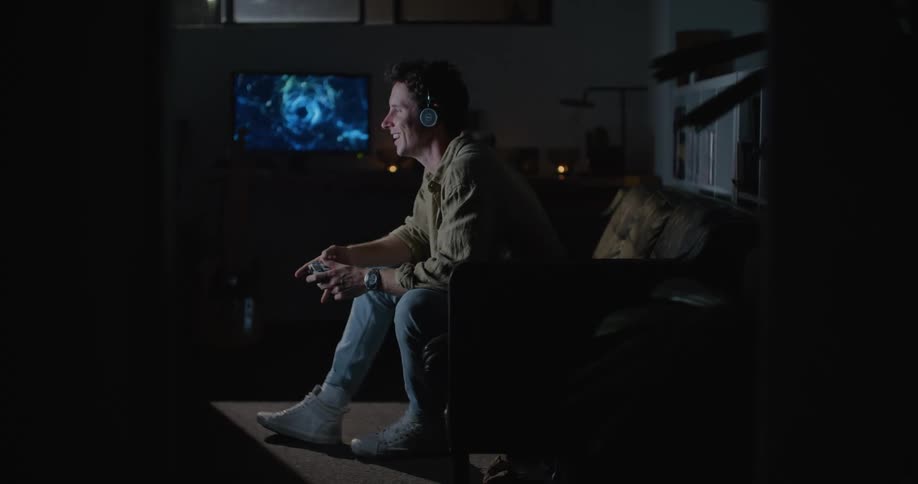 Man wearing a headset sitting at the couch in the living room and playing video game