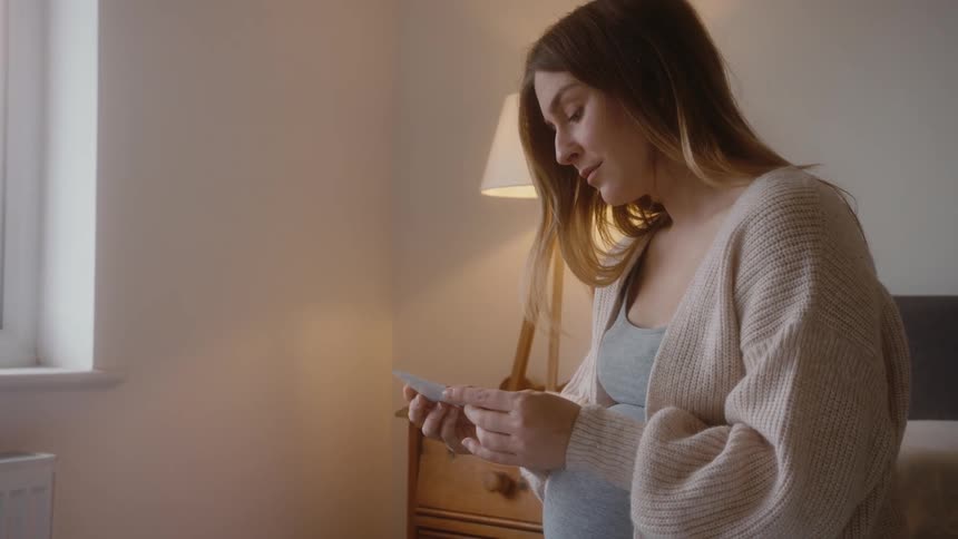 Pregnant woman with brown wavy hair sits on bed and looks at ultrasound in bedroom
