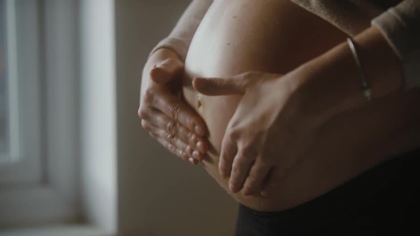 Pregnant woman with beige shirt and black pants rubs her belly