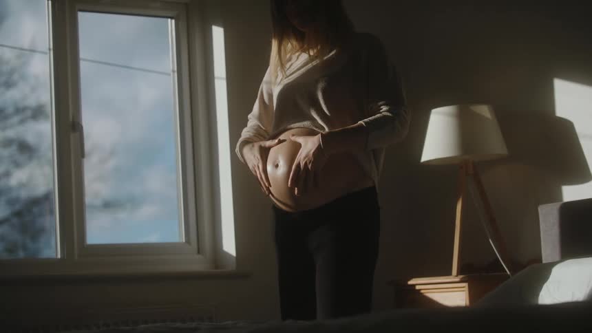 Pregnant woman with grey sweater at home rubs her belly in bedroom