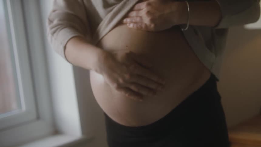 A pregnant woman lifts her shirt to rub her belly while standing near a window indoors during the day