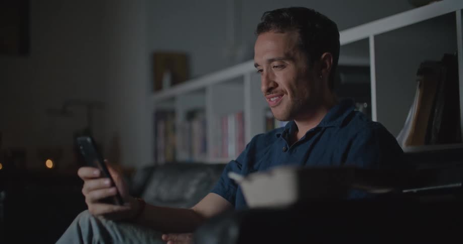 A man sitting on his couch holding his phone reading texts and laughing