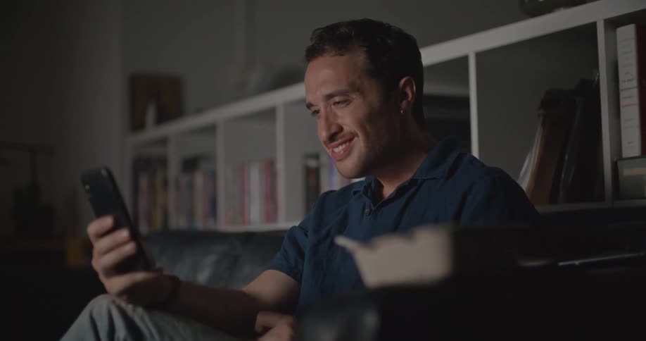 Man with blue shirt sits on sofa looking at smartphone and smiling in living room at home