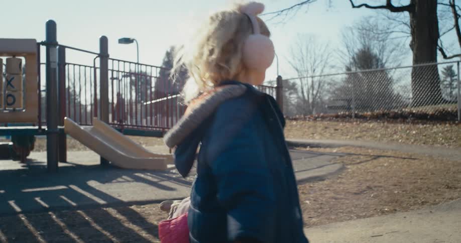 Little blonde girl in the playground outdoors happily running around two other girls that are sitting and playing on the ground at a sunny day