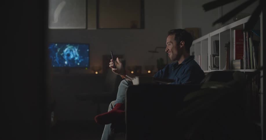 A man uses his smartphone to participate in a video chat while sitting on a sofa in the dimly lit living room of a house