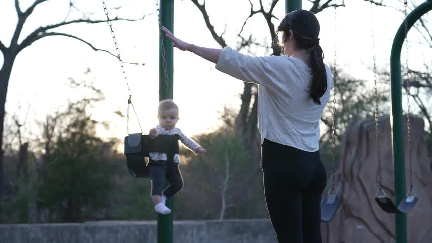 Mother with ponytail white shirt and black pants swings baby in swing seat in playground at sunset