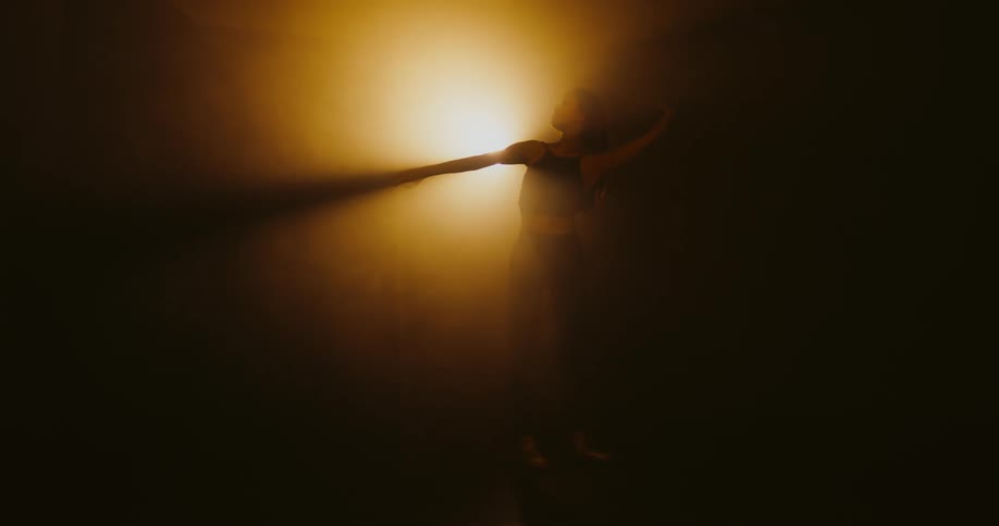A woman dances before a light obscured by mist in a dark place