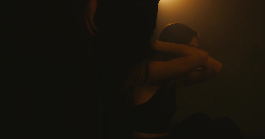 Woman sitting at another persons feet in dim room cover her head with her arms then combs them down her hair