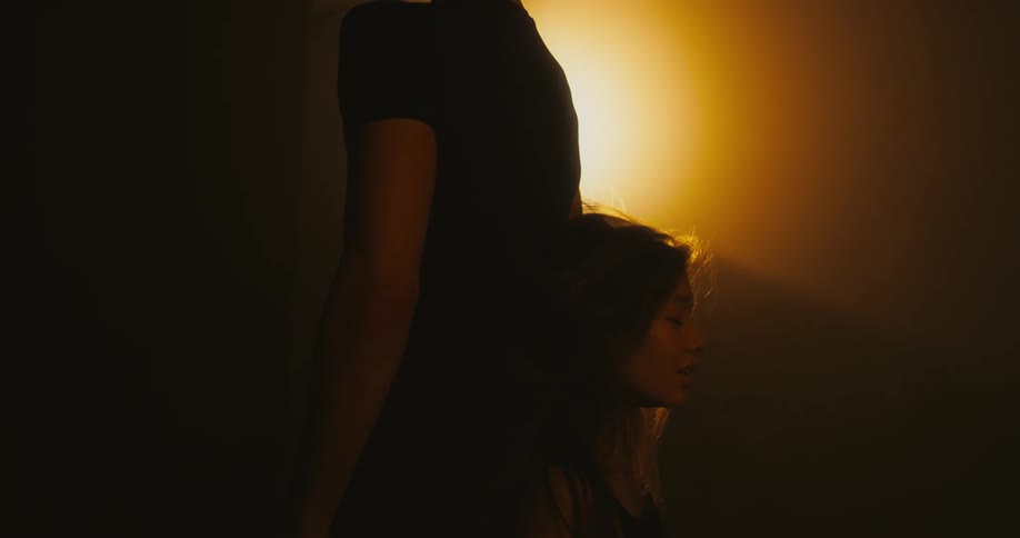 Young woman sliding down the back of a man standing next to her in a dark room with a orange backlight