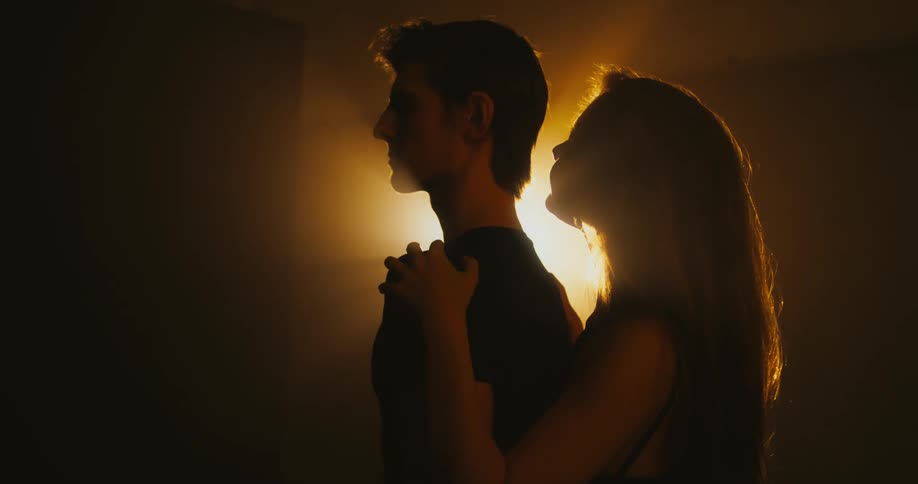 A woman hugs a man from behind as they stand in front of a bright light in a dark place