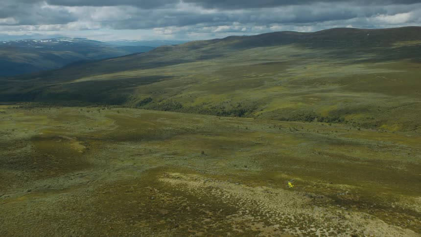A yellow and red rescue helicopter is flying low over hills with mountains in the distance