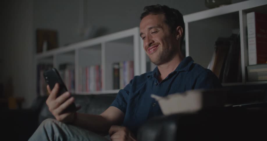 Man with blue shirt and jeans sits on couch using smartphone and making video call living room at home