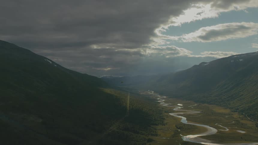 A helicopter flies over a verdant valley between mountains on a cloudy day